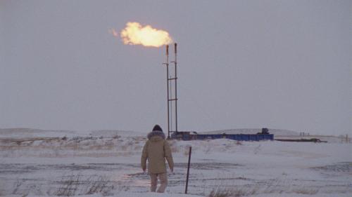 Still from the movie "How to Blow Up A Pipeline." We see the a distant figure from the back as the person watches a burning oil derrick on the far horizon.