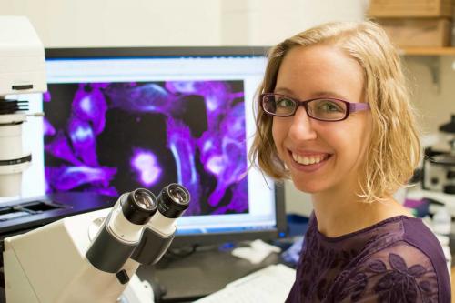 Graduate student in purple shirt and purple glasses posing next to microscope with image of purple microorganisms on a screen in the back